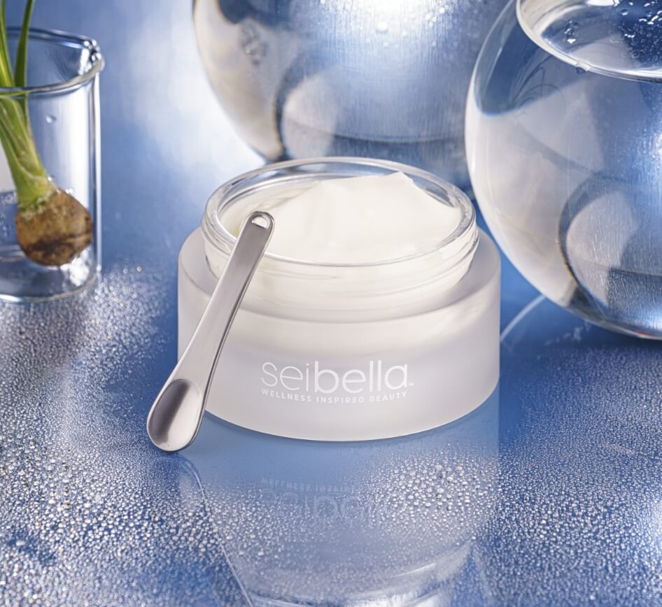 Sei Bella Advanced Epidermal Perfector Cream in an open white jar with a silver spatula, displayed on a reflective blue surface.